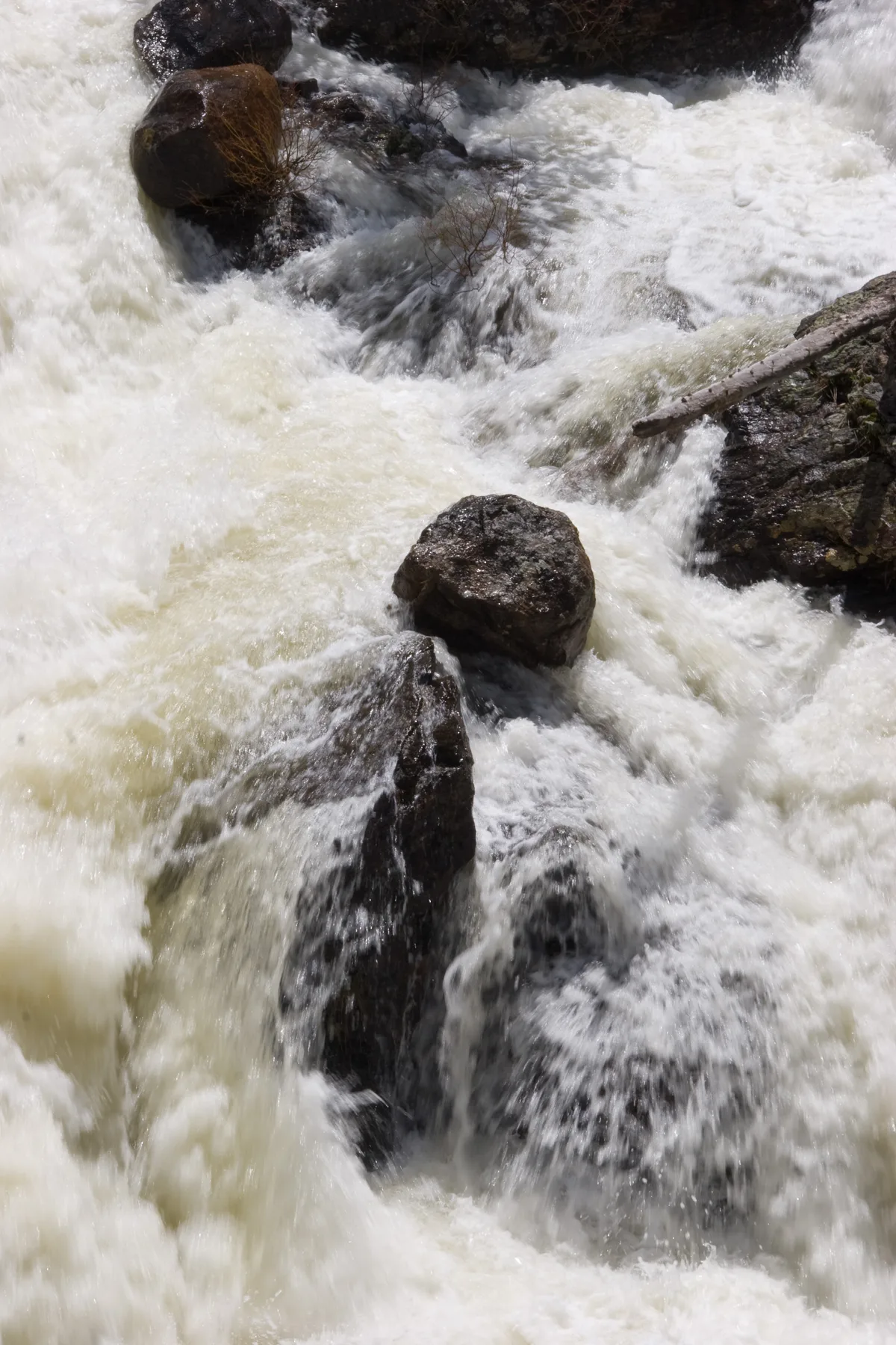 White water surges over dark granite boulders in a tight cascade, with frothy currents splitting around rocks and a small bare branch at the right edge. The close-up composition emphasizes the waterfall’s force and texture rather than the full drop.