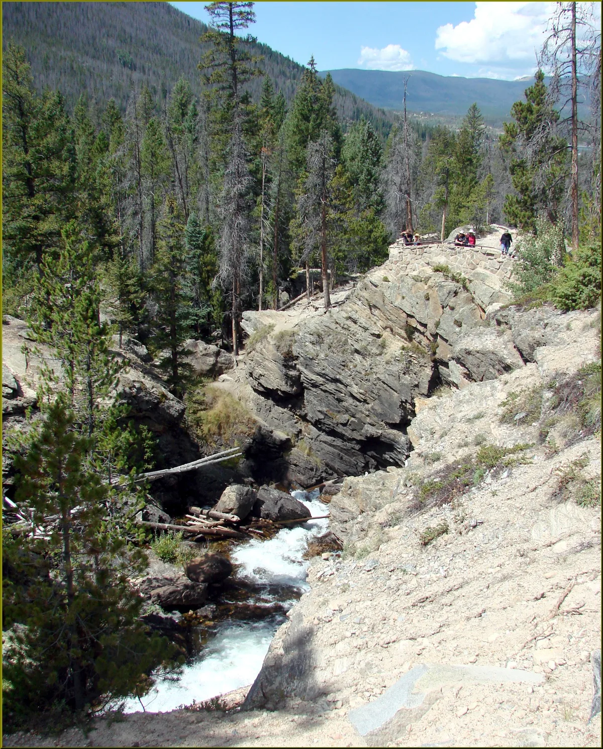 White water rushes through a narrow rocky canyon at Adams Falls, framed by pine trees and sunlit granite ledges. A small stone overlook with a few hikers sits above the falls, with hazy mountain ridges and a bright blue sky in the background.