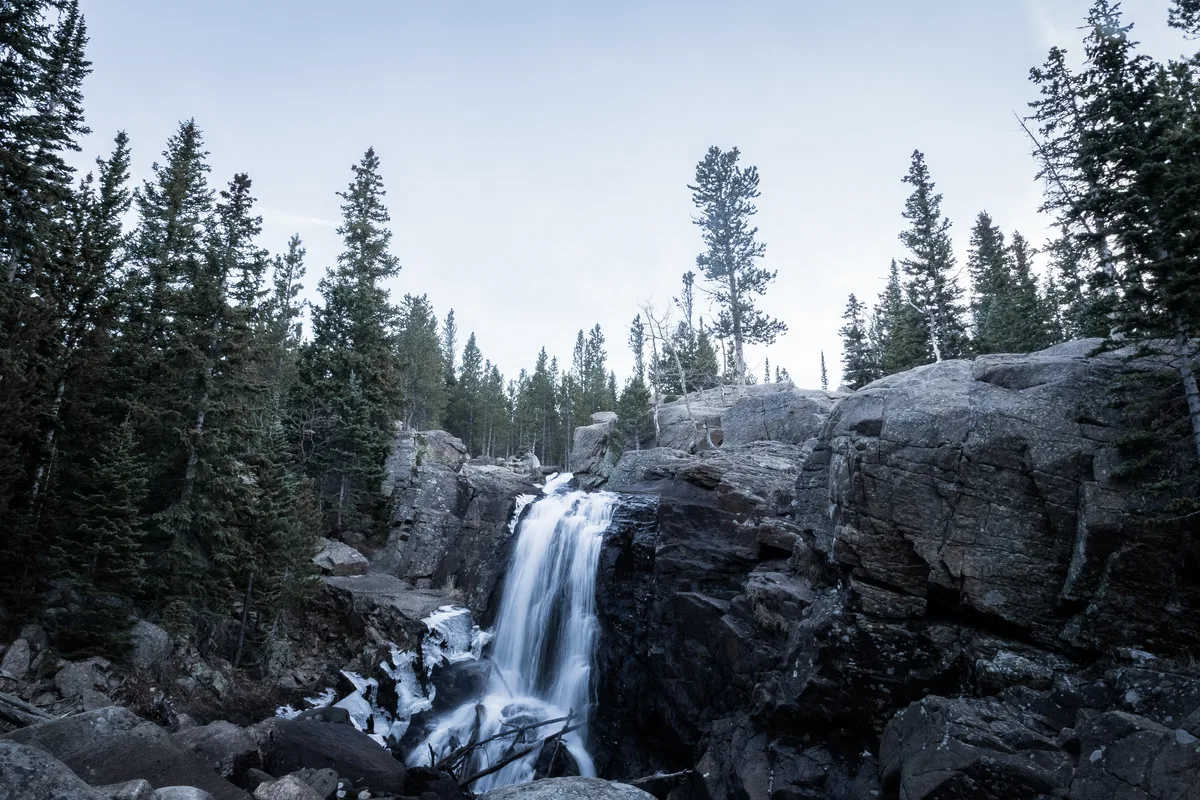 A small waterfall spills through a narrow granite gorge, softened into silky white water by a long exposure as it drops between dark rock walls. Tall spruce and fir trees frame the falls under a pale morning sky, with traces of snow tucked among the rocks.