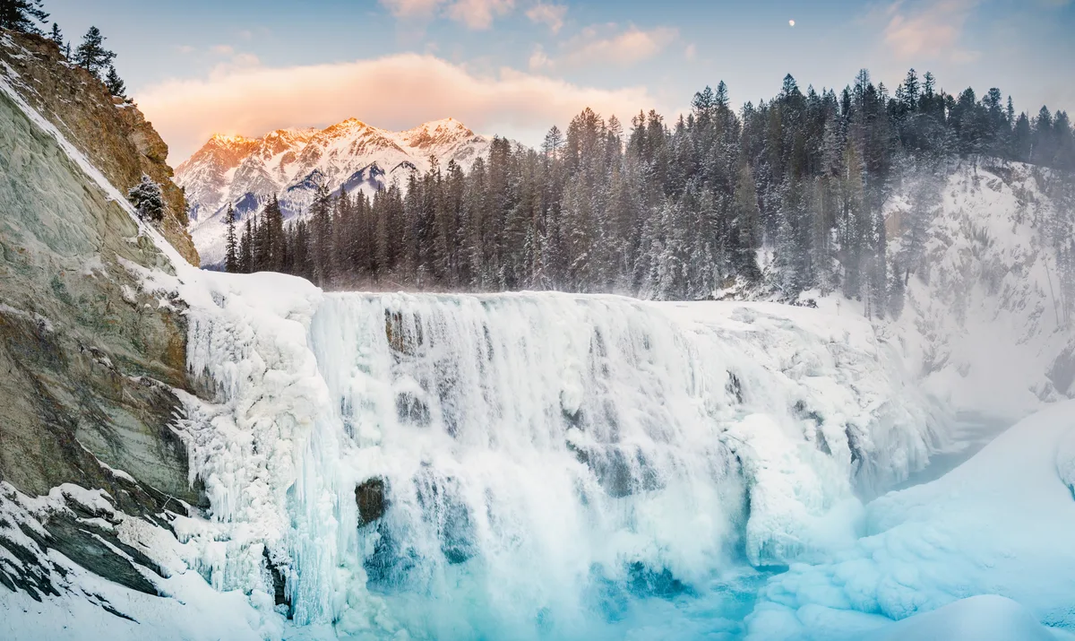 A frozen waterfall spills over a wide, icy ledge into a turquoise pool, with mist rising against snow-covered granite walls. Dark evergreen forest lines the gorge, while warm sunrise light glows on the distant mountain peaks and pastel clouds overhead.