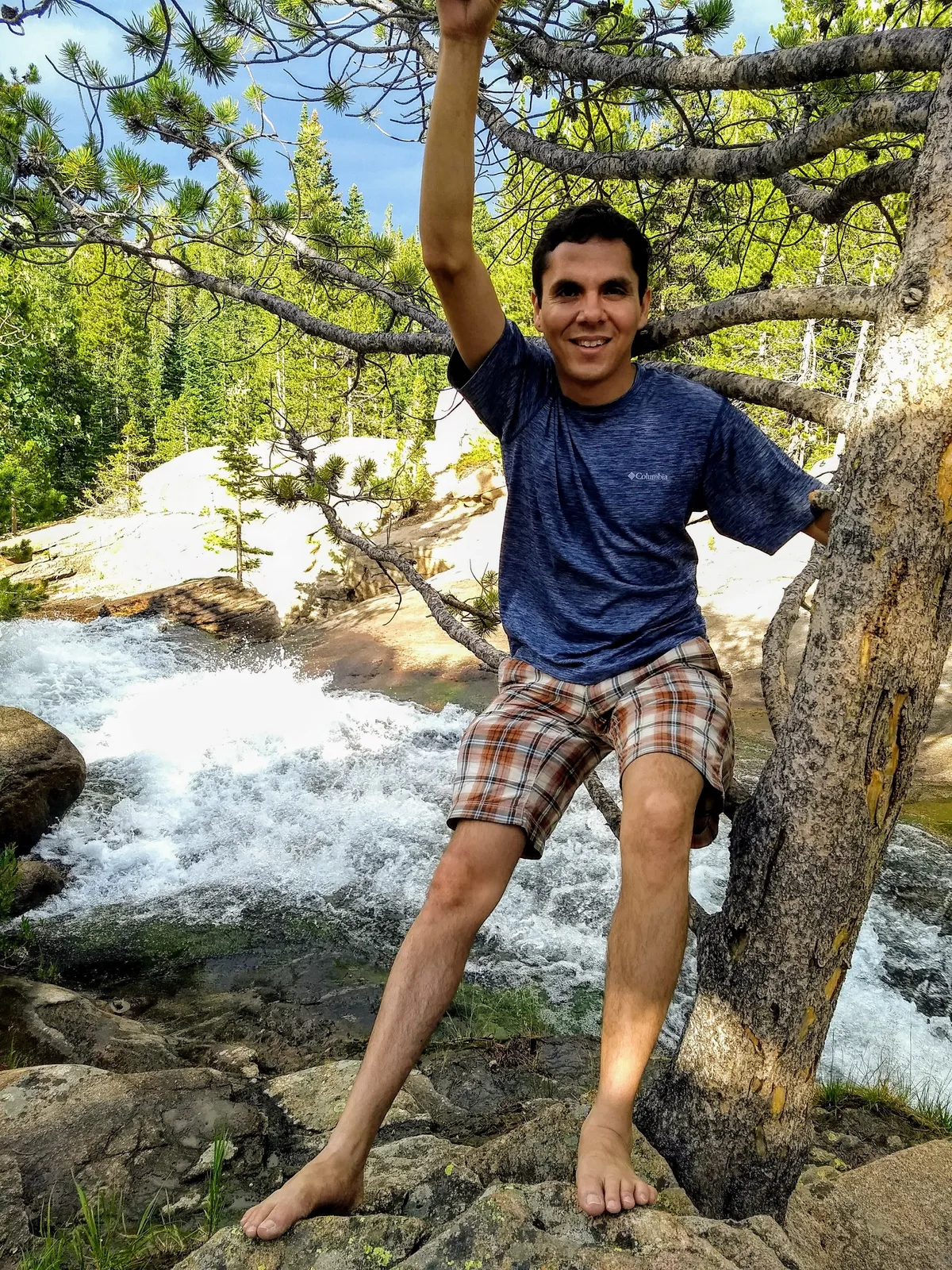 A barefoot man smiles while balancing on rocks beside the rushing water at Alberta Falls, with one arm raised to a pine branch. Sunlit spruce and fir trees frame the cascade, and the white water foams through the granite gorge below.