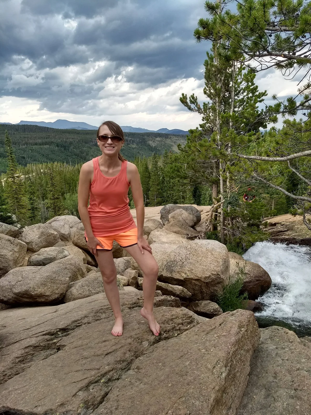 A woman stands barefoot on smooth granite rocks beside a rushing waterfall at Alberta Falls, with evergreen forest and distant mountain ridges stretching under a dramatic cloudy sky. The white water drops through the narrow gorge at right, while the open rocky foreground and bright coral-and-orange outfit draw the eye to her in the scene.