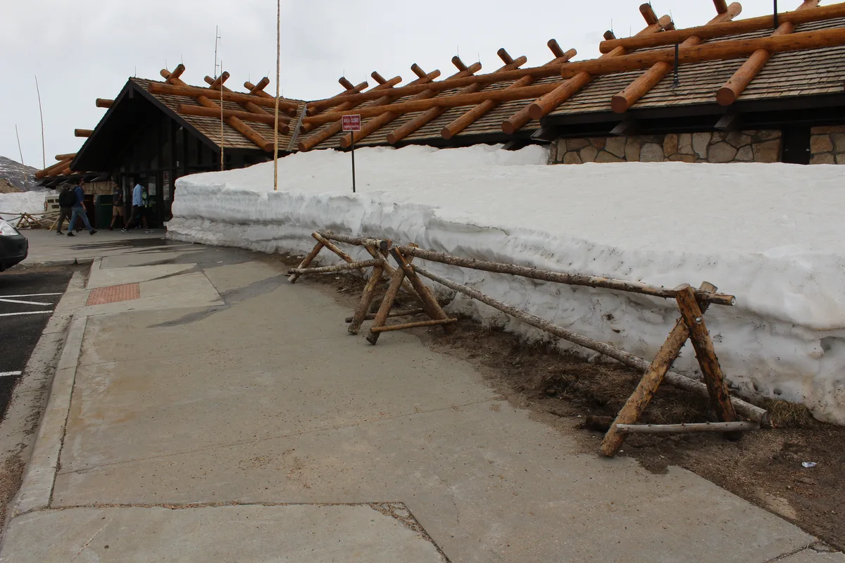 Snowbanks line the walkway outside the Alpine Visitor Center, with a rustic log-roof building rising behind them under a gray morning sky. A few visitors stand near the entrance, while wooden railings and patches of wet pavement emphasize the high-elevation spring thaw.