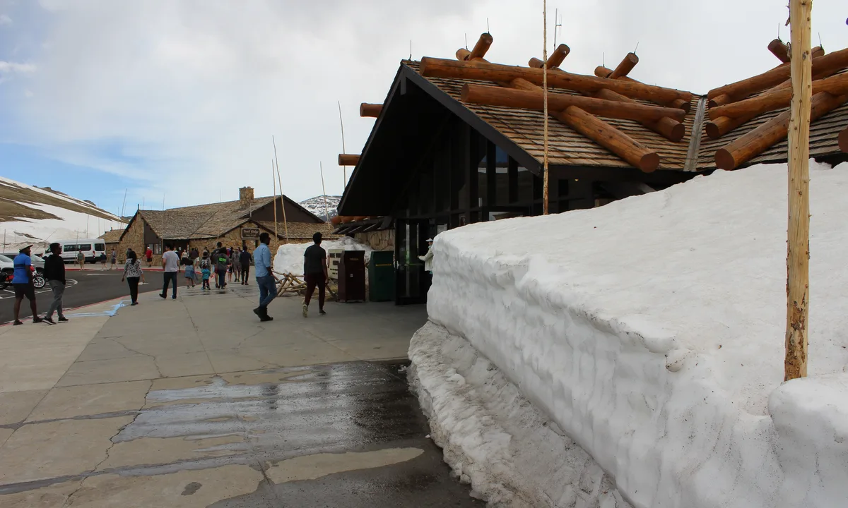 The Alpine Visitor Center sits beside a thick, white wall of lingering snow, with dark log-beam roof details and a wet concrete walkway in the foreground. Visitors walk past the building under a pale morning sky, with snow-covered alpine slopes rising in the background.