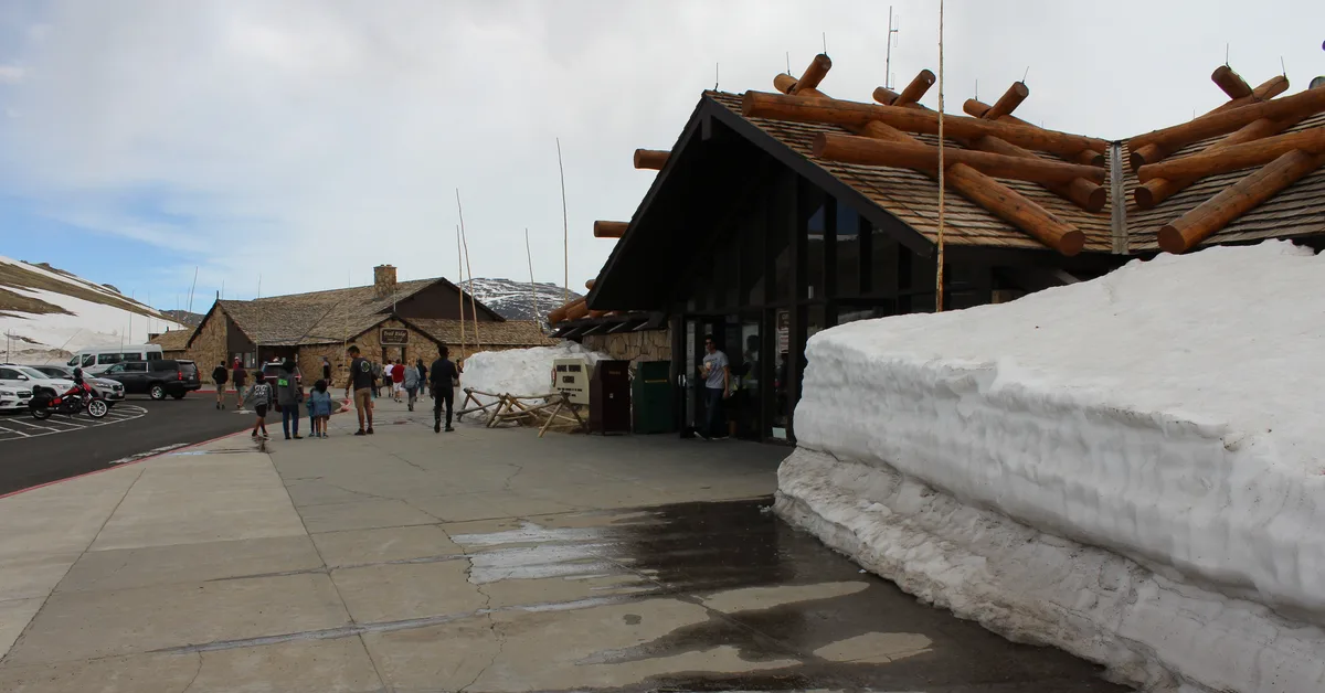Snowbanks line the Alpine Visitor Center plaza, with a wet concrete walkway leading past the log-roofed building and a few visitors walking toward the entrance. Parked cars and a small stone building sit against the high-elevation tundra under a pale morning sky.