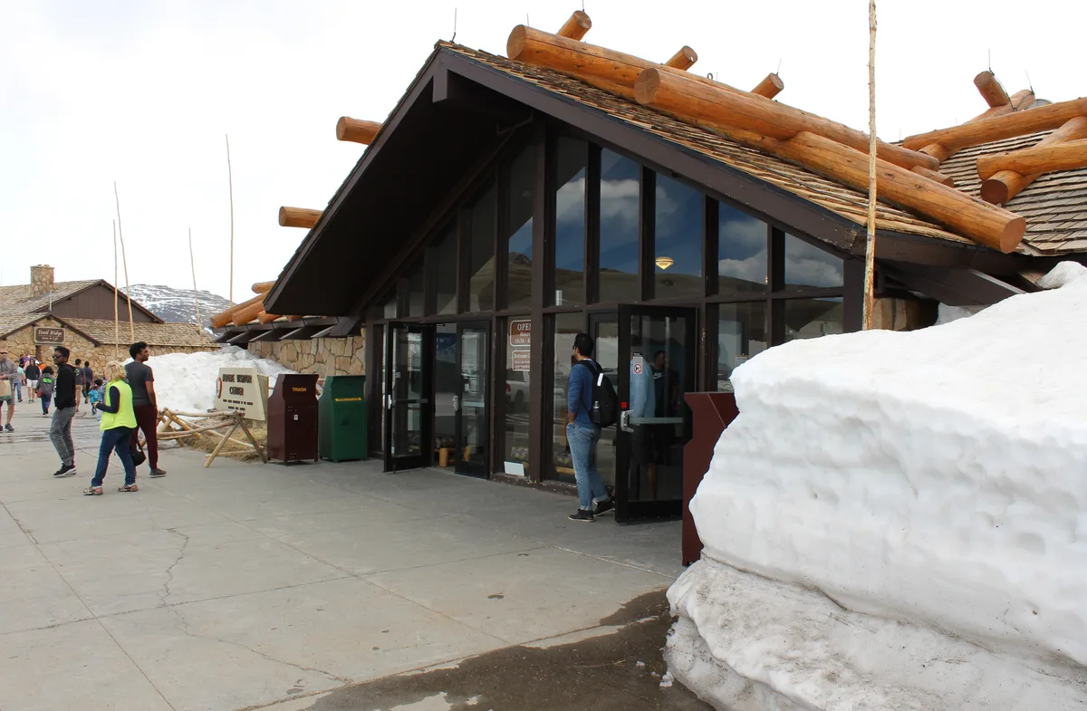 The Alpine Visitor Center’s dark glass entrance and heavy timber roof sit beside tall snowbanks, with visitors walking across the concrete plaza in bright morning light. Stone buildings and a glimpse of snow-covered mountains frame the high-elevation scene.