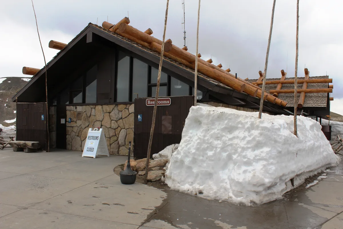 The Alpine Visitor Center sits under a cloudy morning sky, its dark roof trimmed with logs and stone walls facing a concrete walkway. A large snowbank still lingers beside the closed restrooms, with bare poles and wet pavement emphasizing the high-elevation chill.