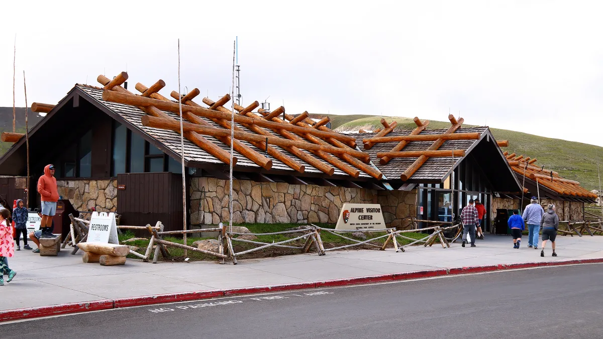Alpine Visitor Center’s low stone building sits beside Trail Ridge Road, with a roof made of exposed log beams and a few visitors walking across the paved entrance in the cool morning light. Green tundra-covered slopes rise behind it under a bright, pale sky.