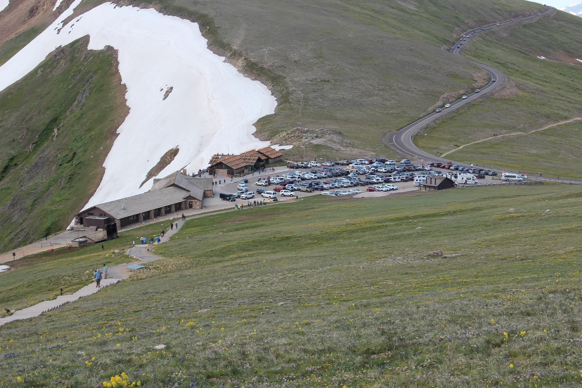 The Alpine Visitor Center sits below a broad snow patch on a high, grassy tundra slope, with a packed parking lot and Trail Ridge Road winding up the mountain behind it. In the foreground, a narrow path crosses the alpine meadow dotted with tiny yellow and purple wildflowers.