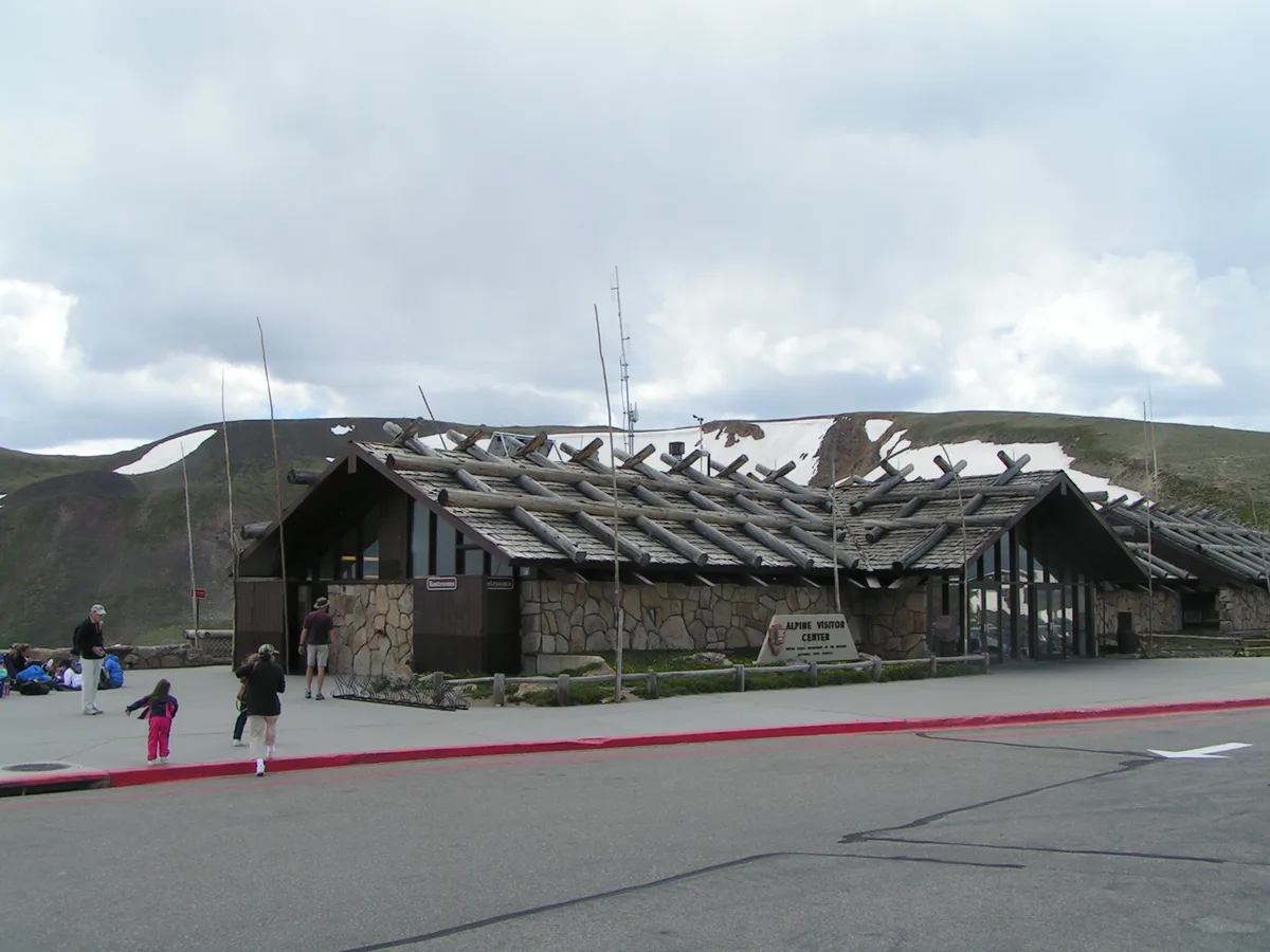The Alpine Visitor Center sits beneath a cloudy sky, its low stone walls and log-beam roof blending into the high-elevation tundra, with patchy snow still lingering on the surrounding hills. A few visitors stand and walk across the paved lot in the foreground.