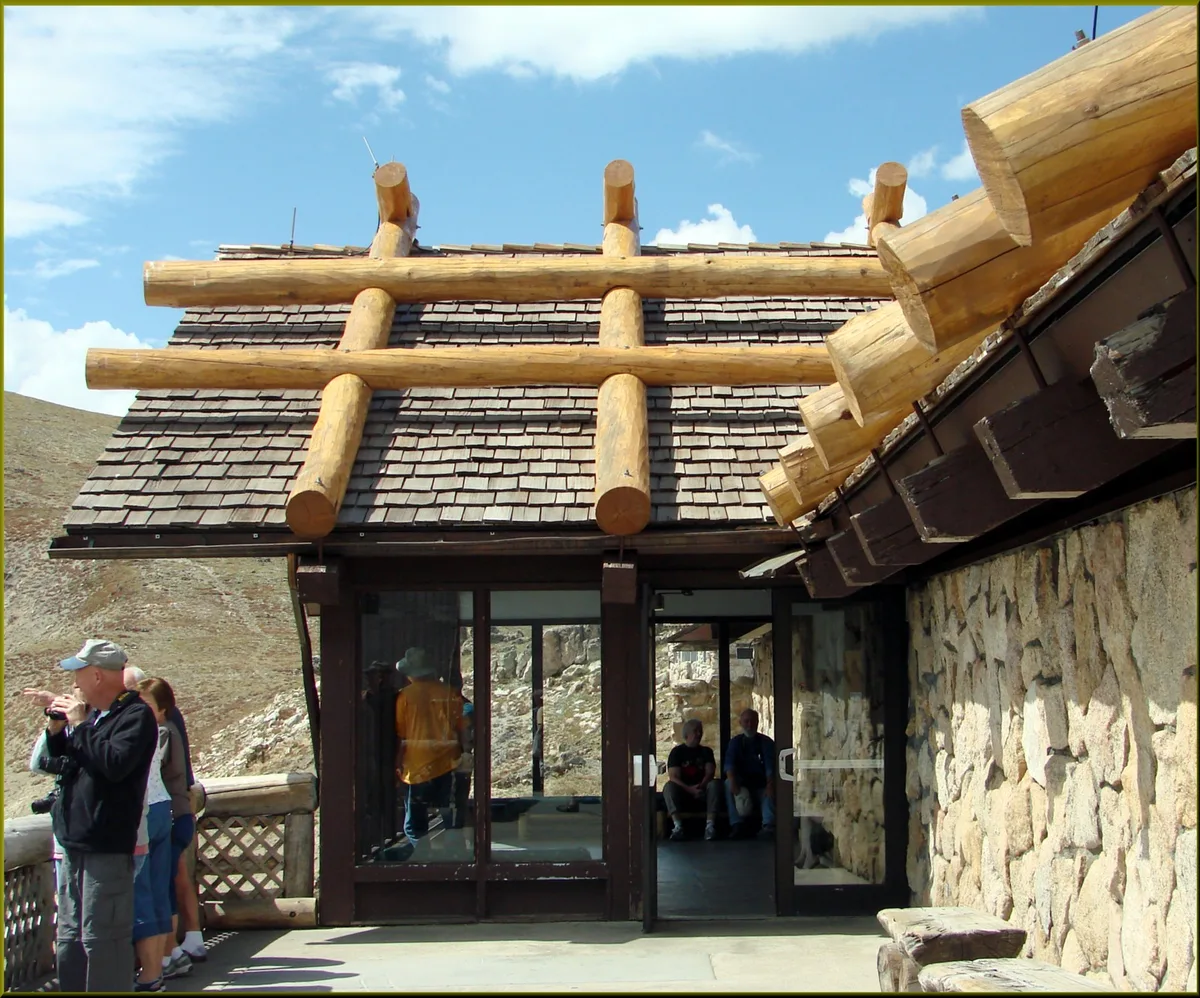 The Alpine Visitor Center’s rustic log-and-stone roof fills the frame, with thick timber beams, shingled slopes, and a glass entrance set against a bright blue morning sky. Visitors stand on the terrace at the edge of the alpine tundra, with barren high-elevation hills rising behind them.