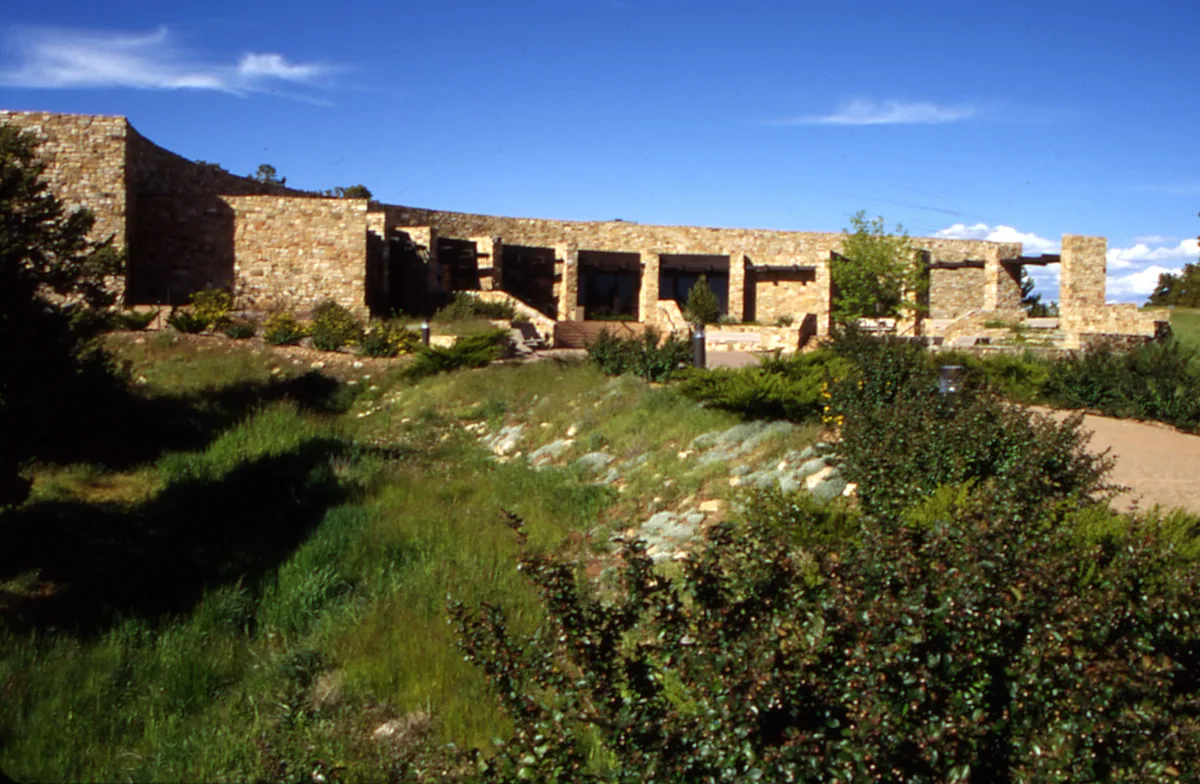 Sunlit stone museum buildings stretch across a grassy foreground, with low shrubs and a curving dirt path leading toward the entrance. The clear blue sky and warm morning light give the scene a bright, high-desert feel.