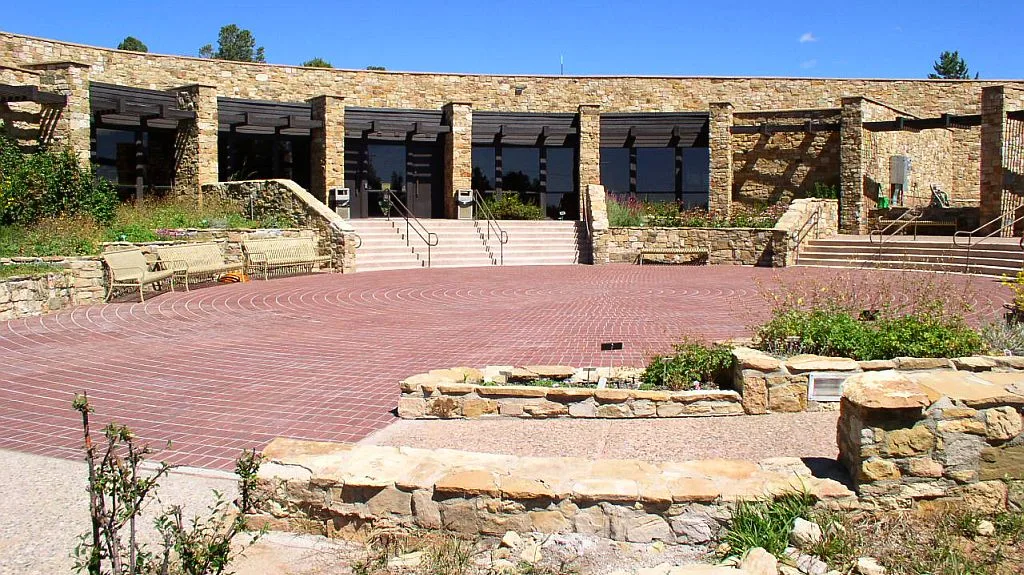 Stone museum buildings curve around a sunlit brick courtyard, with low sandstone walls, steps, and metal railings leading up to dark glass doors. The scene is bright and open, with sparse native plantings and a clear blue morning sky above.