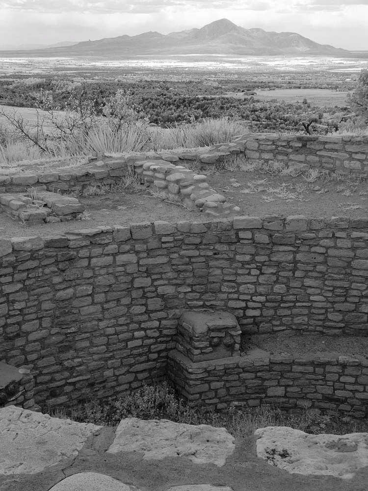 Stone pueblo ruins with curved masonry walls and low room outlines in the foreground, set against a broad mesa landscape and distant mountains under a bright morning sky. The view looks out over scrubby grasses and open country beyond the archaeological site.