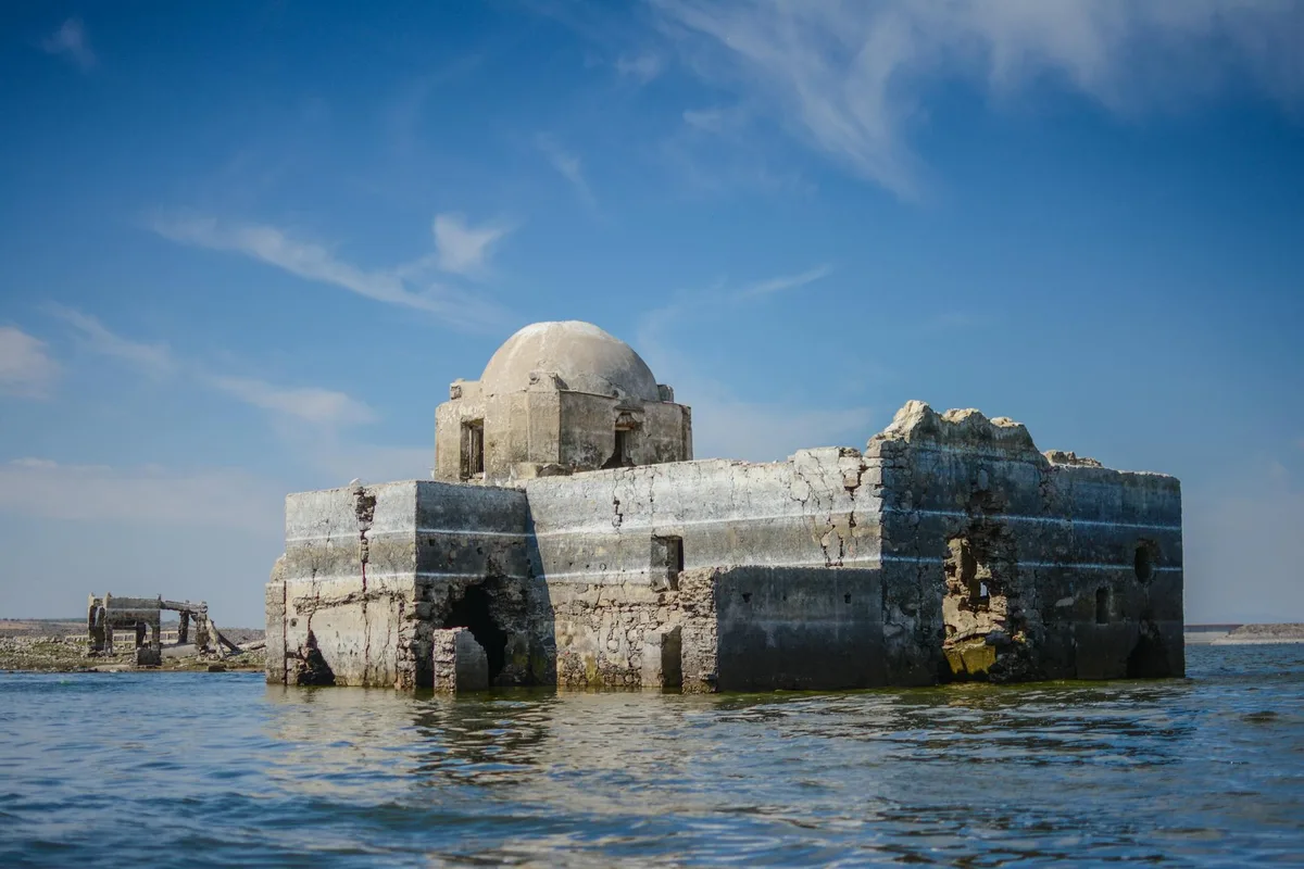 A weathered adobe-like ruin with a rounded white dome rises from blue water, its cracked walls and dark openings catching bright morning light. A smaller broken structure sits to the left under a wide, clear sky.