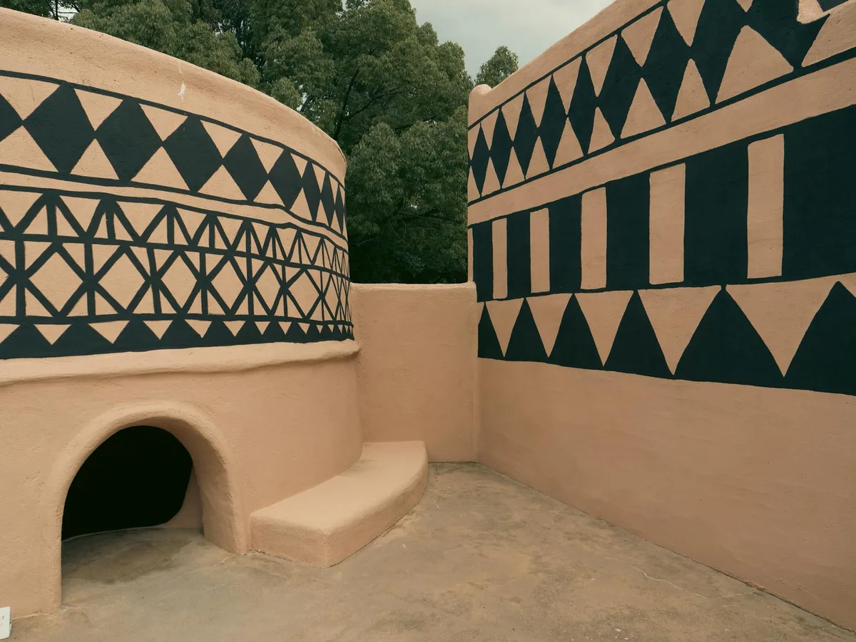 Two tan adobe-style pueblo walls meet in a courtyard, painted with bold black geometric bands of triangles and vertical stripes. An arched doorway and a low curved bench sit in the foreground, with green trees rising behind the structure.