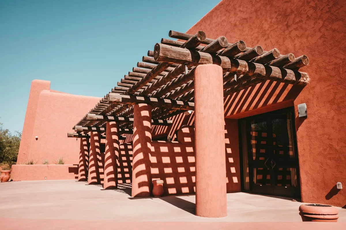 Sunlit adobe-style museum walls and a wooden pergola cast striped shadows across the entrance courtyard. The low, warm-toned buildings sit under a clear blue morning sky, with terracotta planters and clean concrete walkways in the foreground.