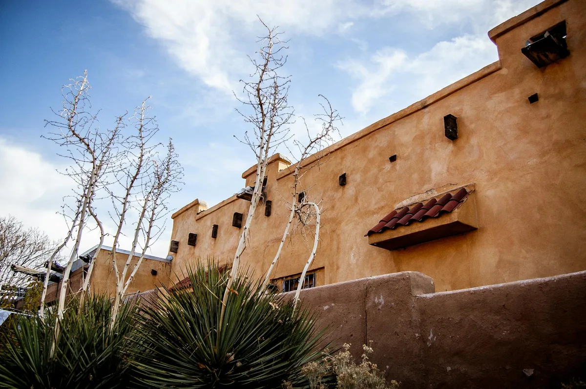 Low-angle view of the Anasazi Heritage Center’s adobe-style wall, with small dark projecting beams and a red-tiled awning under a pale blue morning sky. Leafless desert trees and spiky yucca plants frame the building in the foreground.