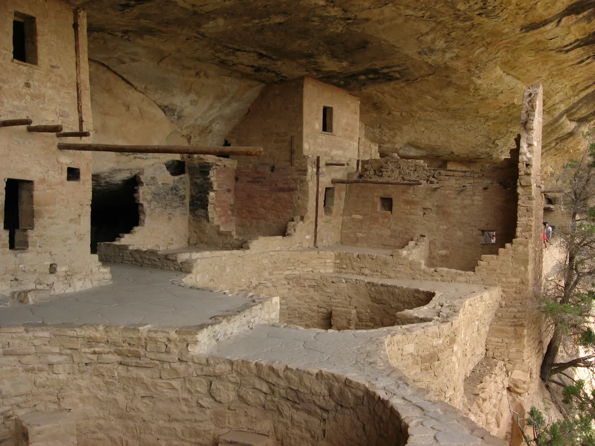 Stone rooms and low walls of Balcony House are tucked beneath a huge sandstone overhang, with dark doorways, wooden support beams, and a curved masonry ledge in the foreground. Morning light warms the tan cliff face while the alcove opens to a narrow view of the canyon edge at right.