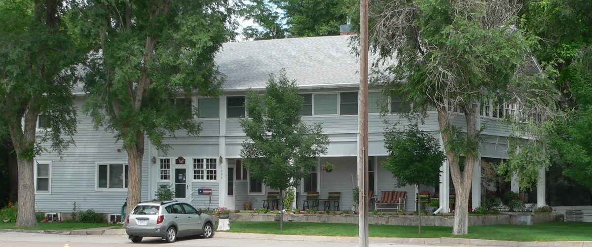 A large white two-story house with a broad covered porch sits behind leafy trees, with a silver SUV parked at the curb in front. The scene is softly lit and suburban, with hanging flower baskets, porch furniture, and a quiet residential street.