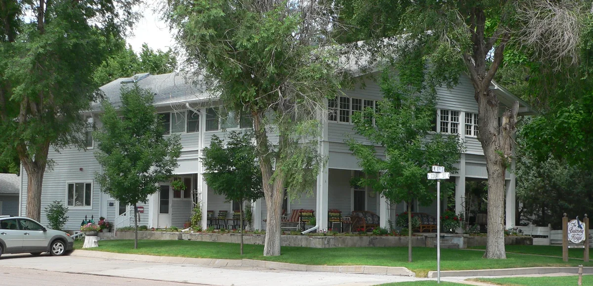 A large gray clapboard house with wraparound porch sits behind leafy trees on a quiet corner, with flower pots, porch chairs, and a street sign in the foreground. The scene is shaded and green, giving it a calm residential feel rather than a cliff-dwelling view.