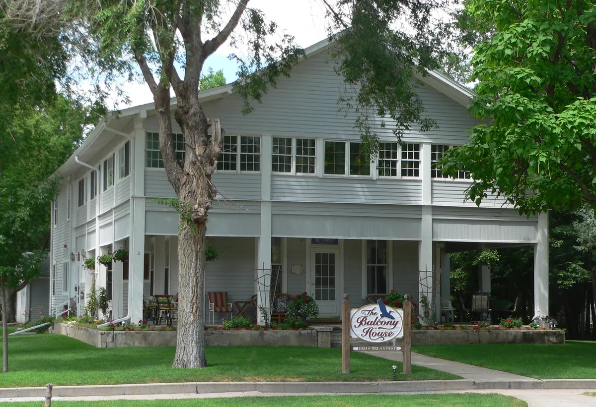 A white two-story house with a wraparound balcony and screened porch sits behind leafy trees, with a sign reading “The Balcony House” in the front yard. The scene is bright and green, with flower boxes, a trimmed lawn, and a sidewalk leading to the entrance.
