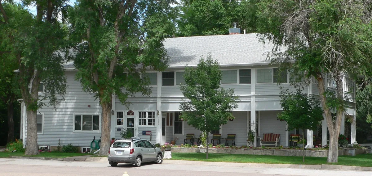 A broad gray two-story house with a wraparound porch sits behind mature trees, with a silver SUV parked at the curb in front. White trim, porch chairs, and flower beds soften the facade in the morning light.