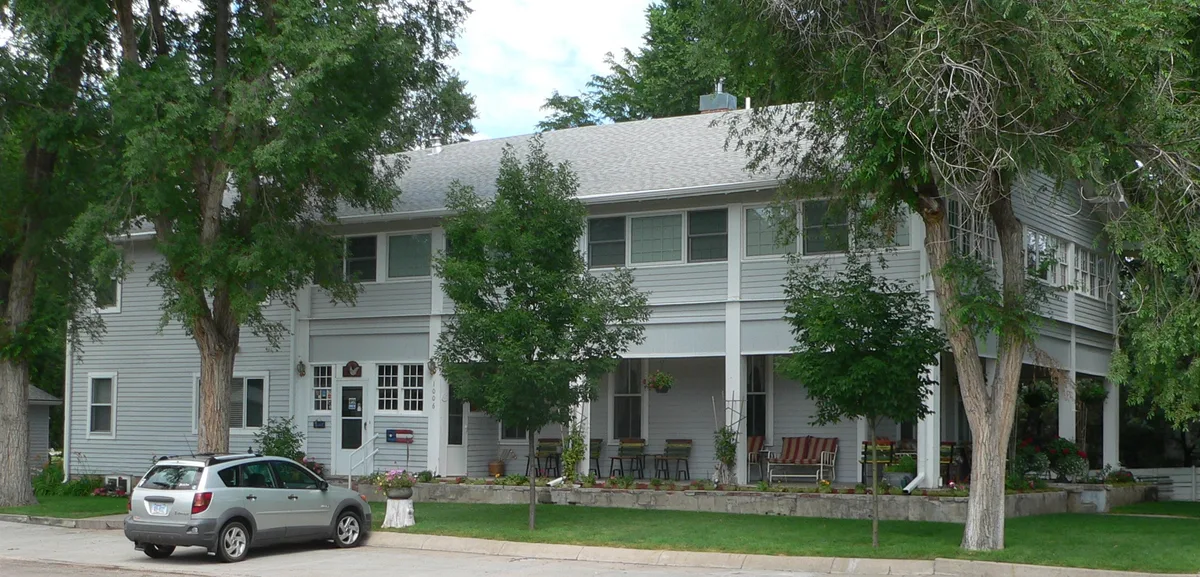 A two-story white clapboard house with a long shaded porch sits behind tall trees, with a silver SUV parked at the curb in front. The scene is bright and quiet, with neatly kept grass, porch chairs, and hanging flower baskets.