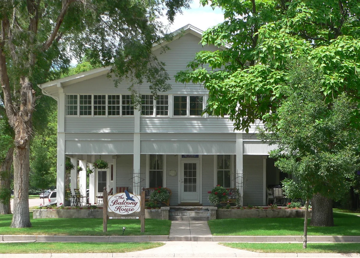 A white two-story house with a broad front porch and rows of windows sits behind a shaded green lawn, framed by leafy trees. A sign in front reads “The Balcony House,” with flower pots and porch chairs adding a welcoming, bed-and-breakfast feel.