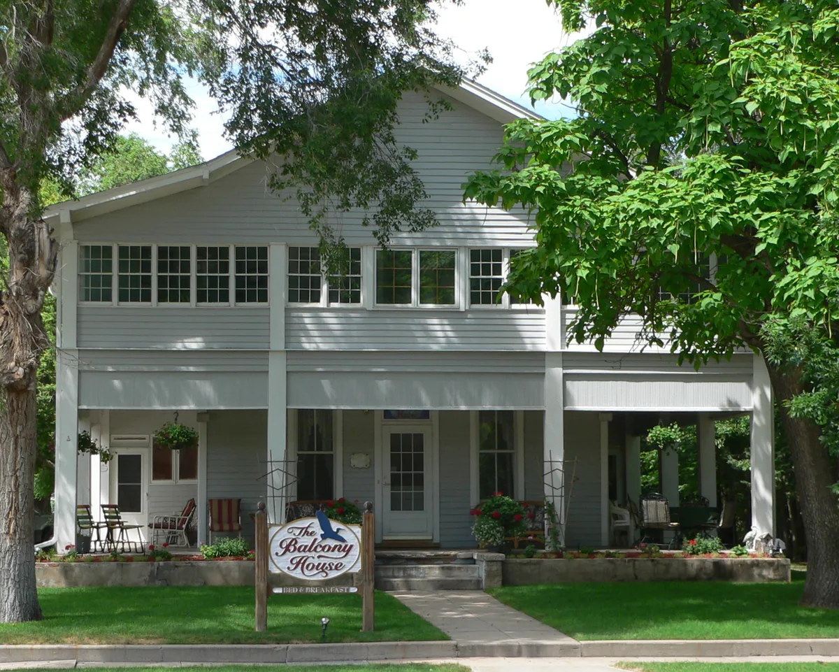 A two-story gray bed-and-breakfast with a deep front porch sits behind leafy trees, with white trim, rows of windows, hanging flower baskets, and a central sign reading “The Balcony House.” Sunlight and shade dapple the lawn and facade, giving the home a quiet, garden-like feel.