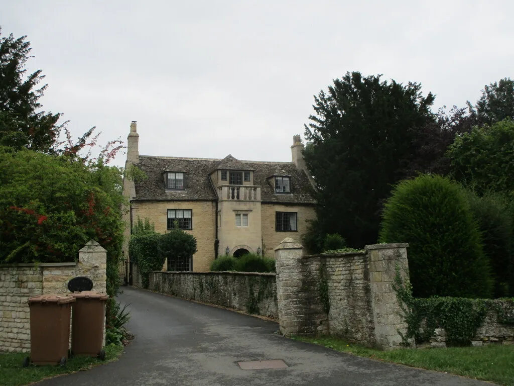 A narrow driveway curves up to a stone house with a steep shingled roof, central balcony, and small-paned windows, partly hidden by dense trees and hedges. Low stone walls and gateposts frame the entrance under a soft gray morning sky.