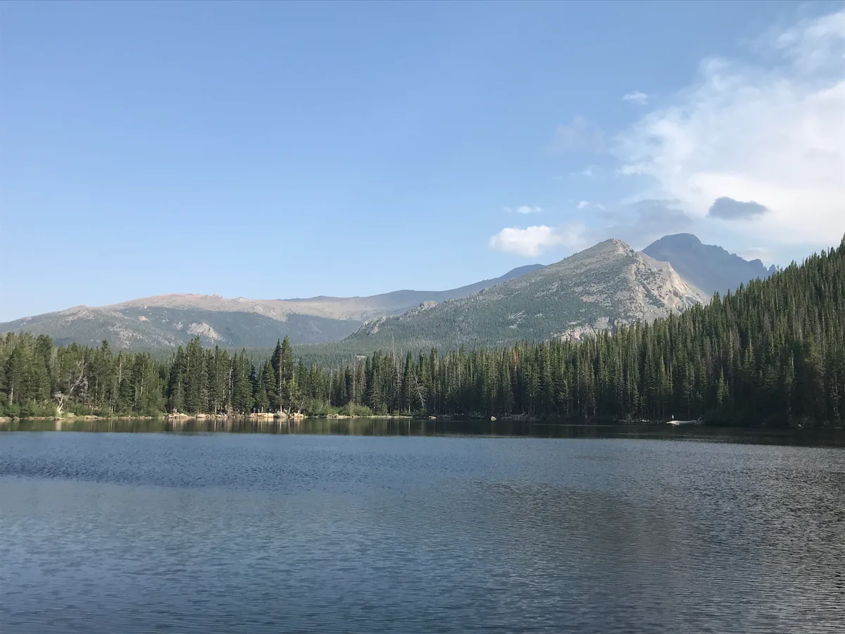 A calm alpine lake fills the foreground, reflecting a dense evergreen shoreline and rugged mountain peaks under a clear blue sky. Sunlit slopes and a soft cloud bank frame the high-country scene.
