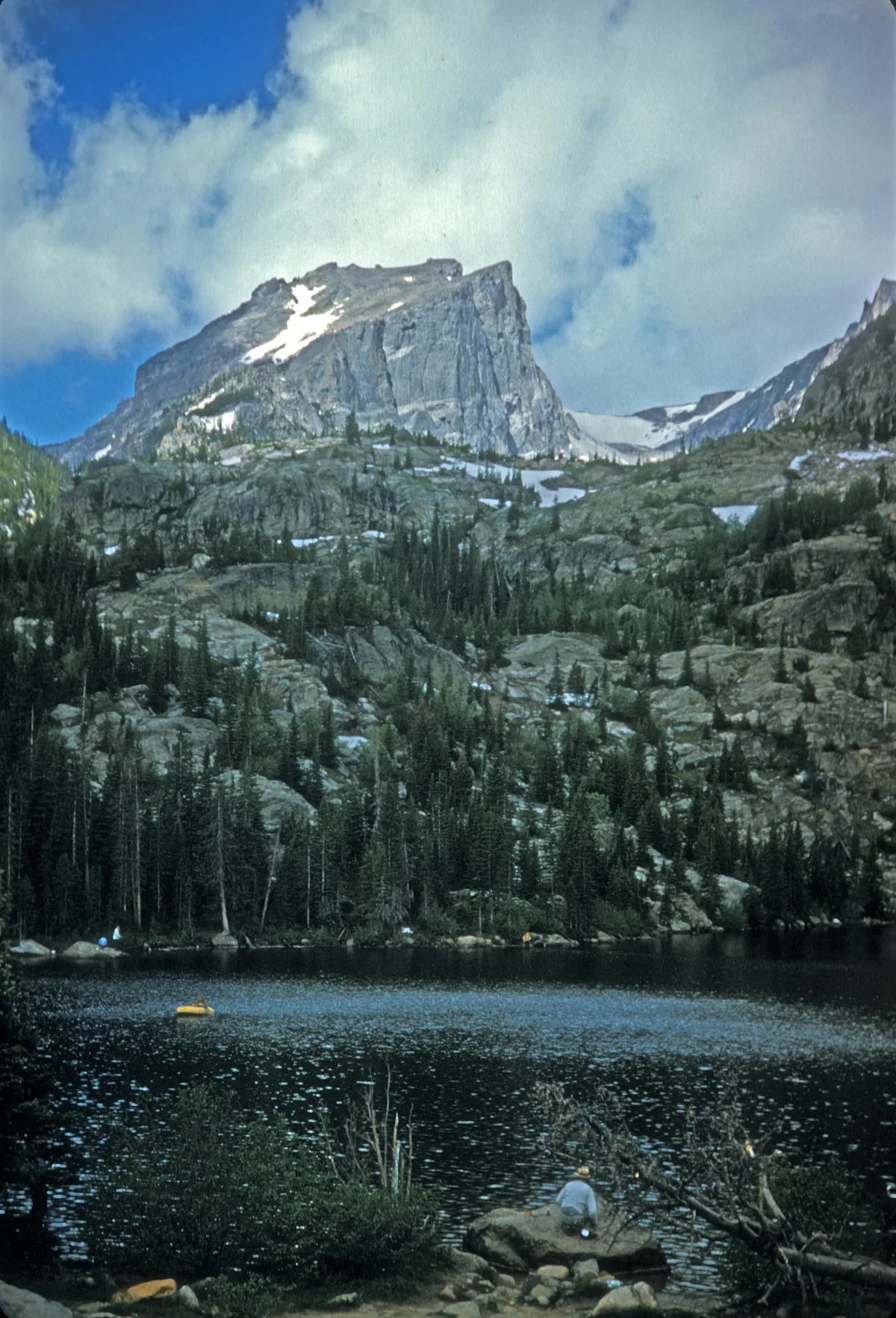 Bear Lake sits below a massive gray granite peak with patches of lingering snow, framed by dark evergreens and rocky slopes. The calm, dark water fills the foreground, with a few hikers and a small yellow kayak adding scale beneath a bright, cloud-streaked sky.