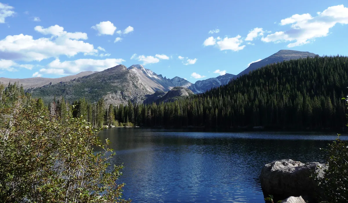 Calm Bear Lake fills the foreground with deep blue water, framed by dark evergreen slopes and rugged granite peaks under a bright blue sky with scattered clouds. The scene feels crisp and open, with the mountain ridges and forest reflected faintly in the lake.