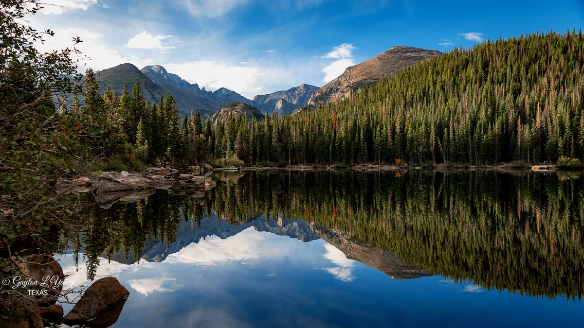 Calm Bear Lake mirrors the surrounding evergreen slopes and rugged peaks in glassy morning water, with blue sky and a few clouds reflected nearly perfectly below. Rocky shoreline boulders and dense alpine forest frame the wide view of the Continental Divide mountains.