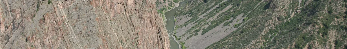 Sheer, streaked canyon walls drop into a razor-thin gorge, with the Gunnison River threading through the bottom far below. The wide view emphasizes the massive scale of the dark precambrian rock and the tight, vertigo-inducing gap between the cliffs.