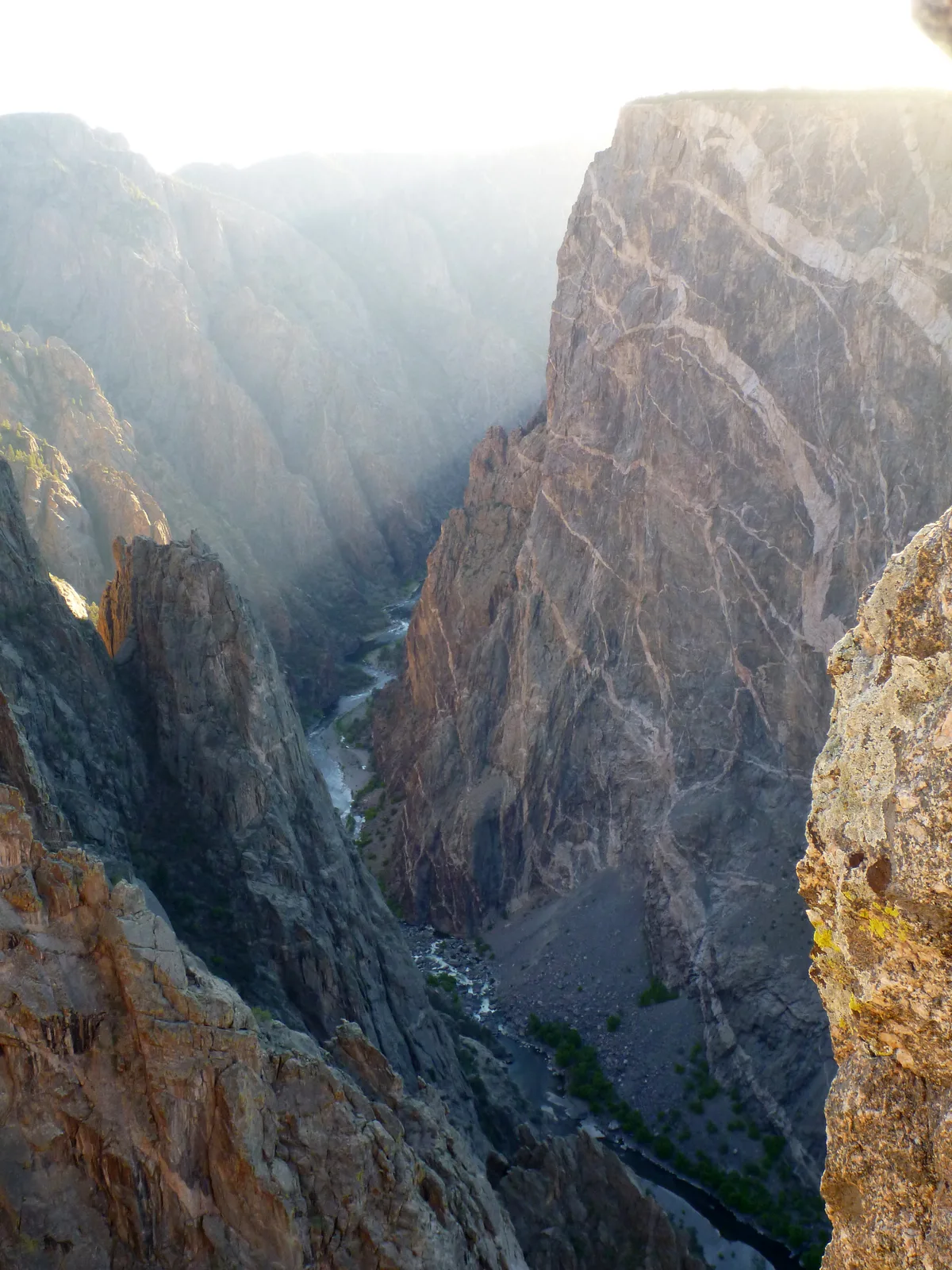 Morning light washes over the sheer dark walls of Black Canyon of the Gunnison, with the Painted Wall rising on the right in pale streaks of pink and white pegmatite. Far below, the Gunnison River threads through the narrow canyon between steep rock faces and shadowed slopes.