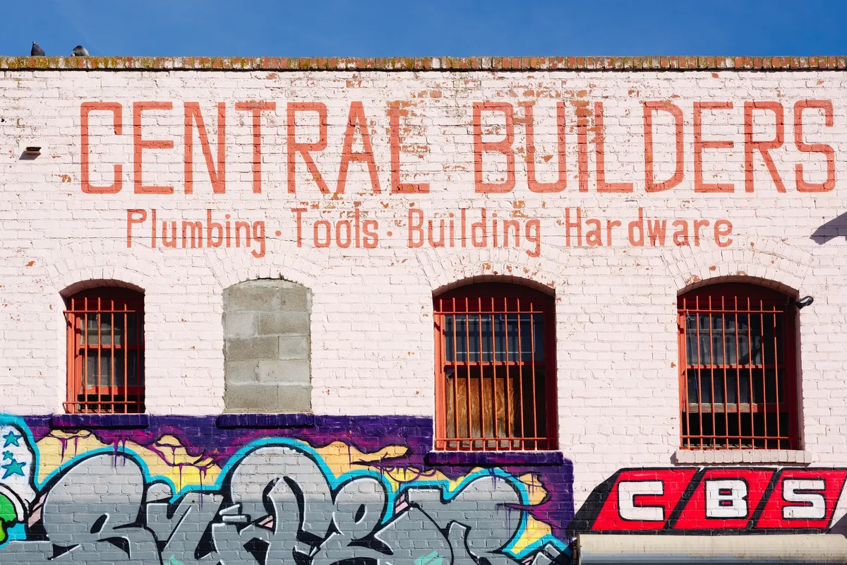 A weathered white brick building fills the frame, with faded orange painted signage reading “CENTRALE BUILDERS” above barred windows. Bright graffiti bands across the lower wall add bold purple, blue, and red accents against the sunlit facade.