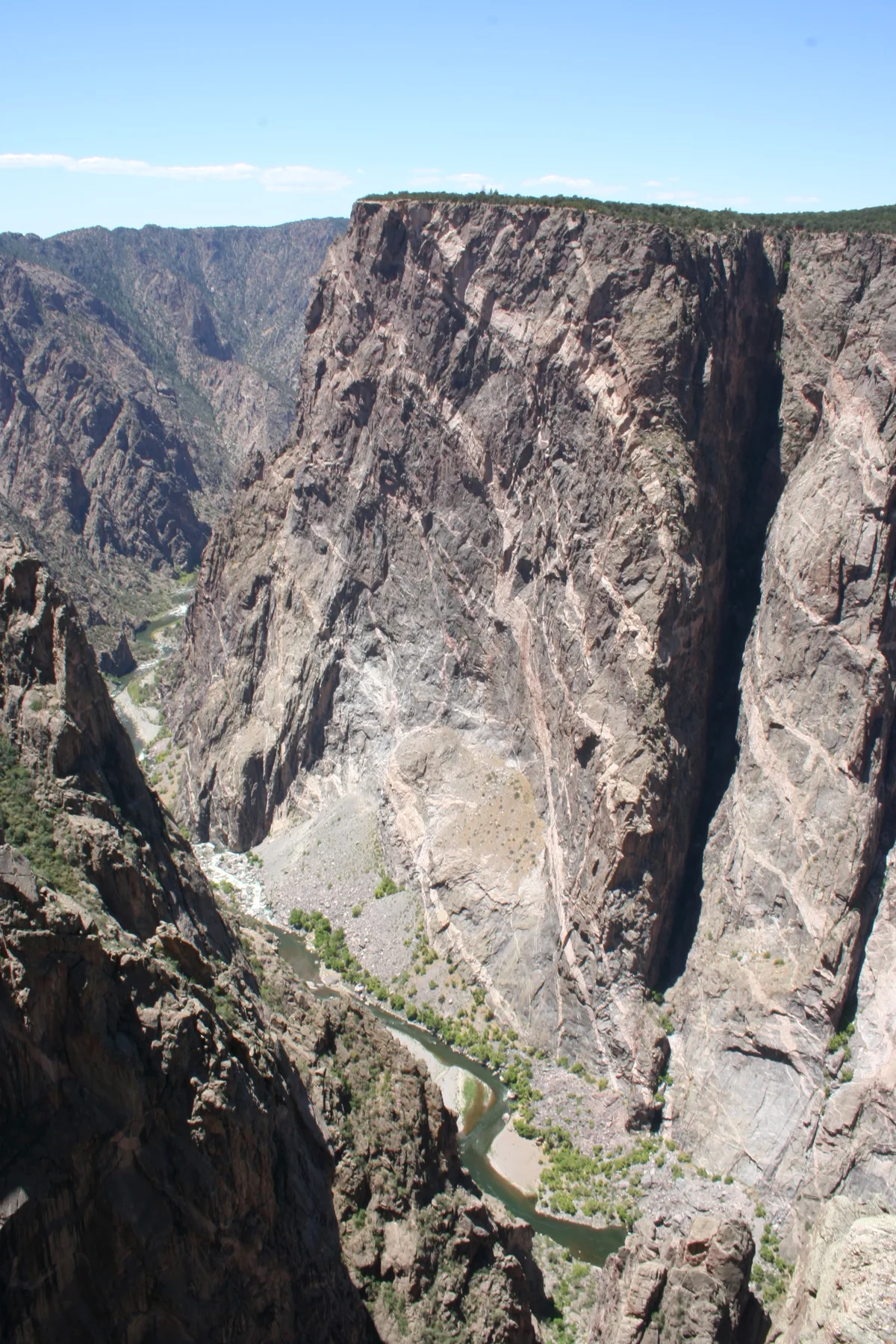 A towering dark granite cliff fills the frame, streaked with pale pink and white pegmatite veins and dropping steeply into the narrow Black Canyon below. The Gunnison River winds through the bottom of the gorge under a clear blue morning sky.