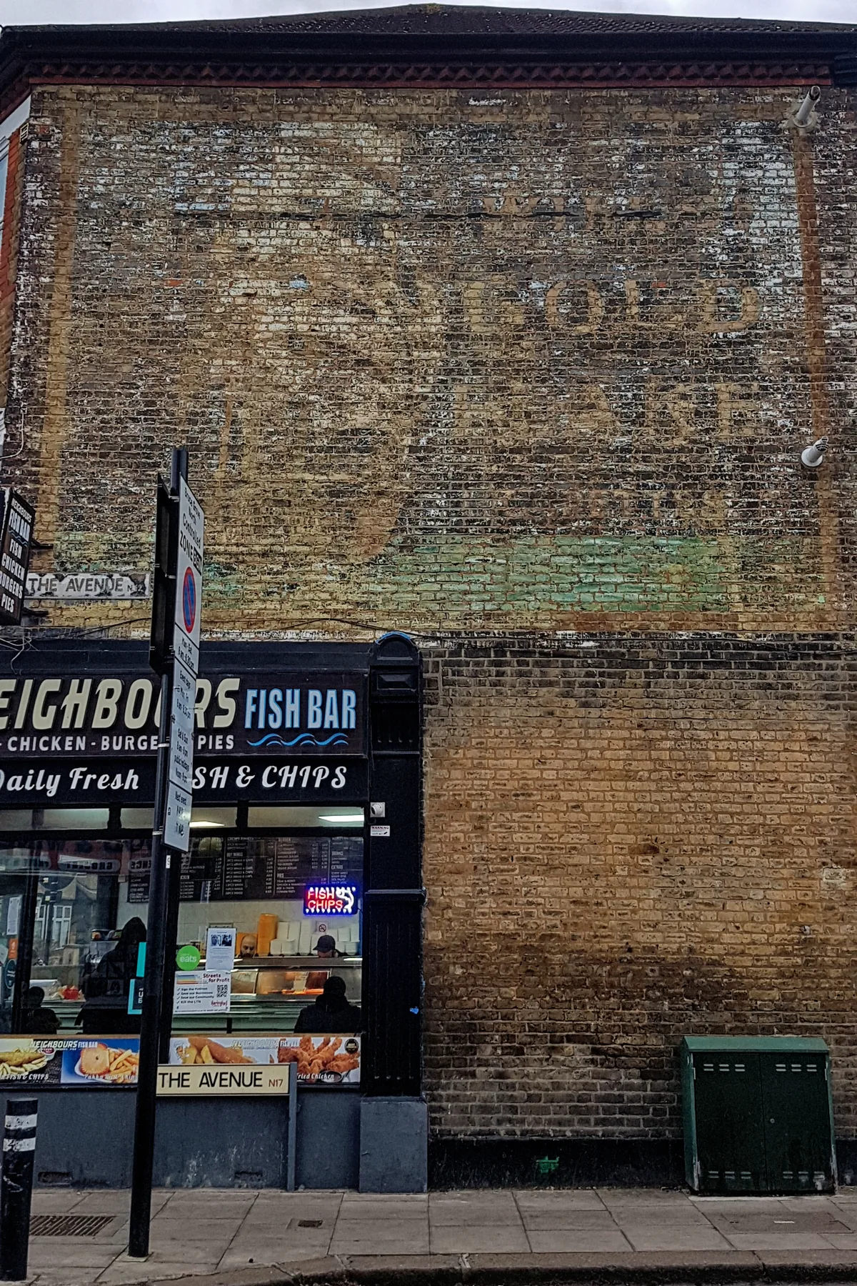 A weathered brick wall fills the frame, with a faded ghost sign barely visible across the upper facade. At street level, a fish and chips shop sits beneath the old lettering, with a glowing neon “FISH CHIPS” sign and a sidewalk signpost in front.