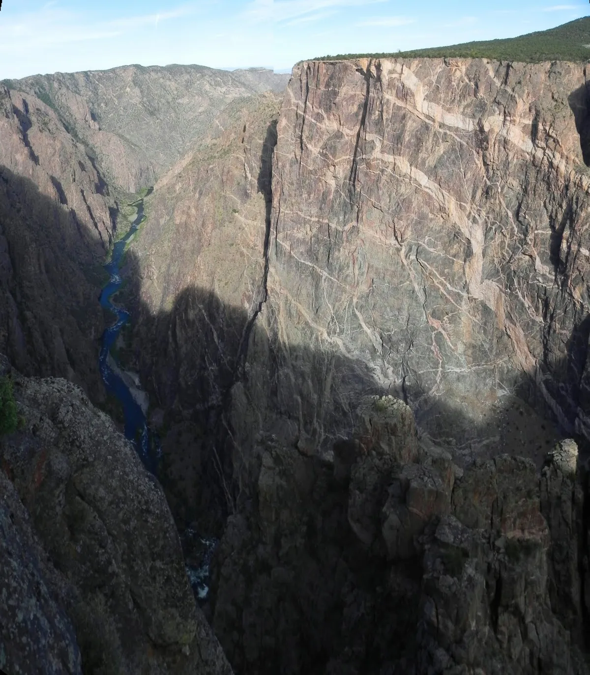 Sheer black canyon walls drop to a narrow blue-green river far below, with the Painted Wall dominating the right side of the frame and streaked by pale pink and white pegmatite veins. Morning light highlights the upper cliff while deep shadows carve out the canyon’s steep depth.