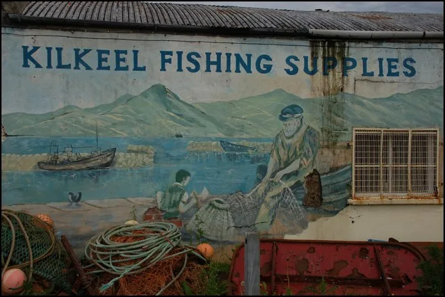 A faded mural on a weathered building reads “KILKEEL FISHING SUPPLIES,” with painted boats, fishermen, and pale mountains stretching across the wall. Coiled ropes, fishing gear, and a barred window sit below the mural, adding to the worn harbor feel.