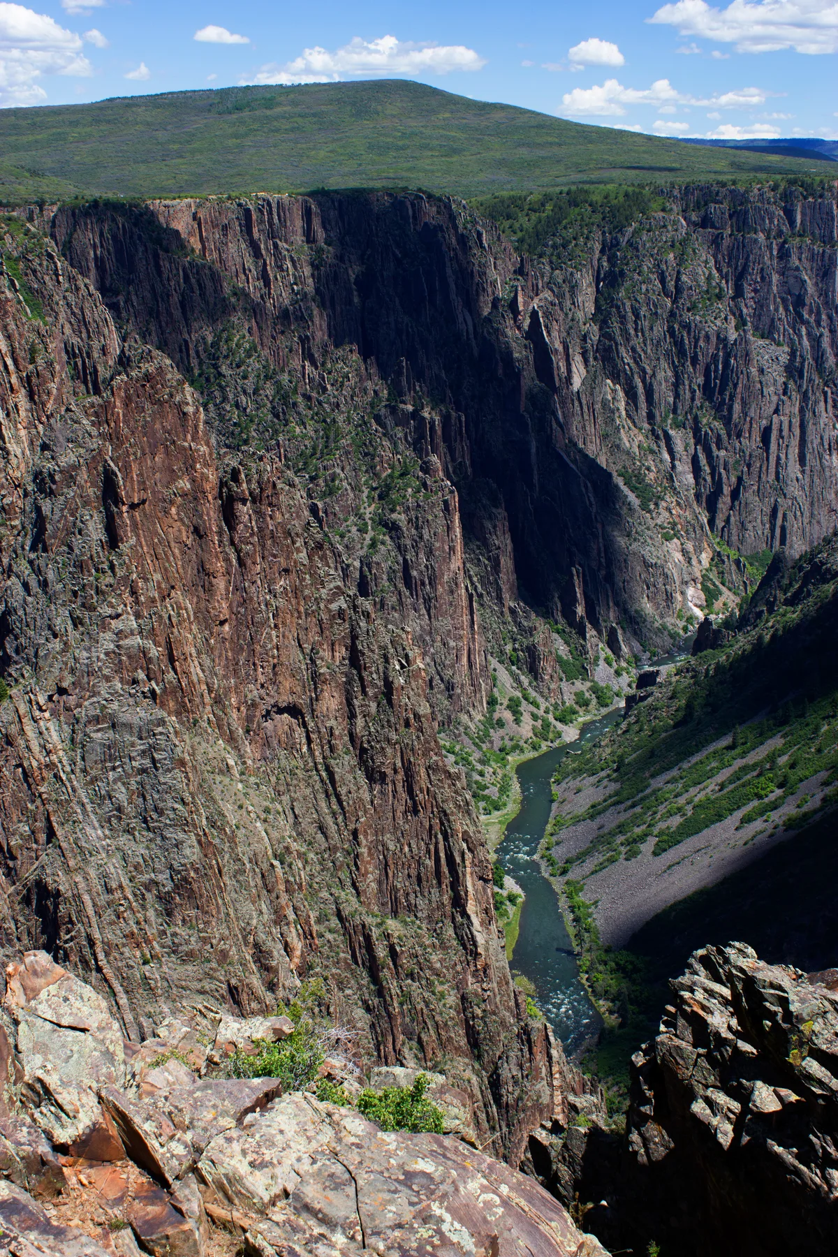 Steep, striated canyon walls drop dramatically to the Gunnison River, which winds through the narrow gorge below. Bright sunlight hits the rust-colored rock and green slopes under a blue sky with scattered clouds, while the lower canyon lies in deep shadow.