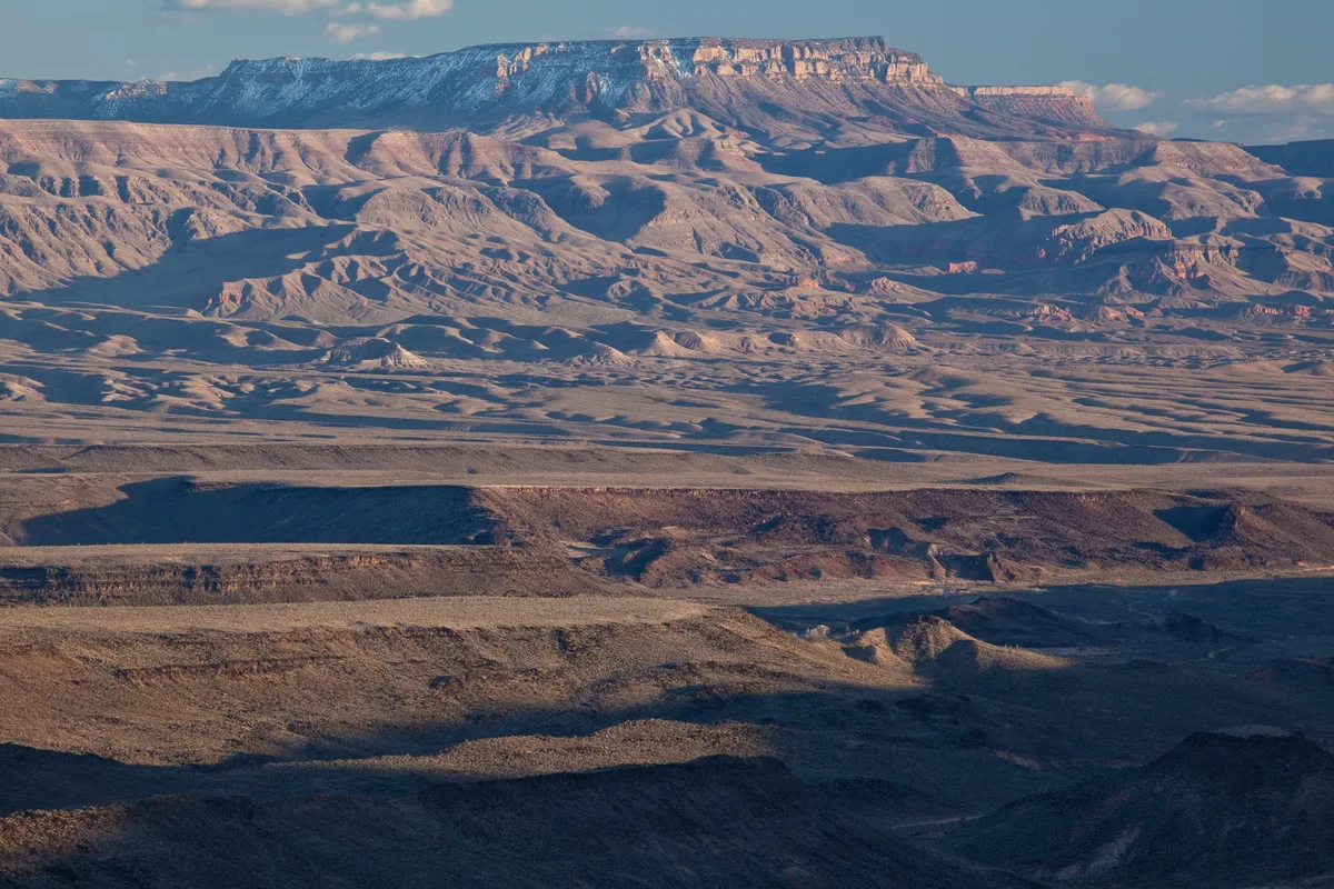 Layered desert mesas and rugged badlands stretch into the distance, with a flat-topped plateau catching warm morning light under a clear blue sky. Long shadows carve across the sandy ridges, emphasizing the remote, eroded landscape.