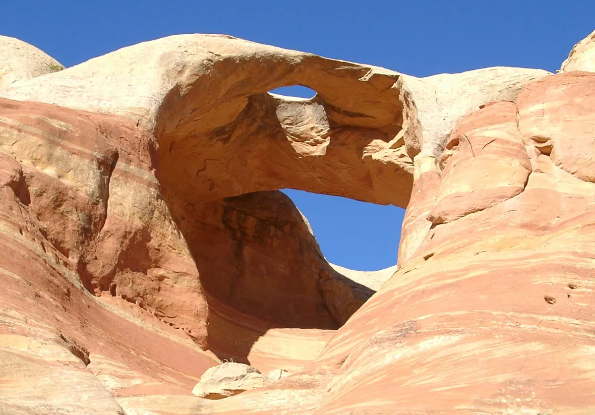 A natural sandstone arch fills the frame, its smooth orange-and-cream rock glowing in bright sunlight against a deep blue desert sky. Layered, wind-carved walls and a smaller opening beneath the arch emphasize the rugged shapes of Rattlesnake Canyon.