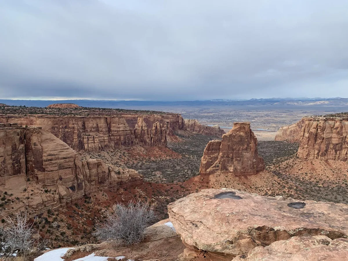 A wide view over a rugged desert canyon with towering red sandstone cliffs, a solitary butte in the center, and a distant valley stretching to low mountains under a heavy gray morning sky. The foreground shows a flat rock ledge with sparse shrubs and a few patches of snow, emphasizing the remote winter setting.