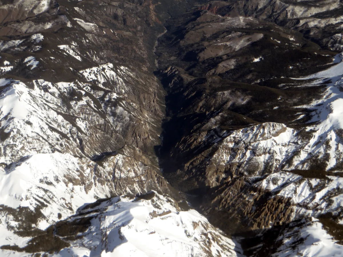 Steep, snow-streaked alpine ridges frame a narrow dark gorge cutting through the San Juan Mountains. Bright morning light picks out the white slopes and rugged rock textures, emphasizing the dramatic depth of the valley.