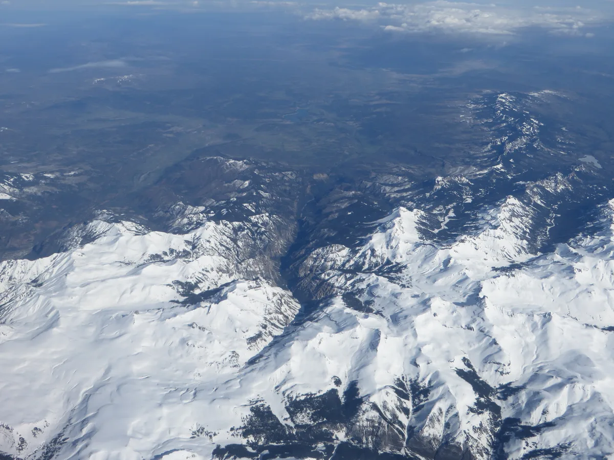 Snow-covered mountain ridges and dark forested valleys stretch across a vast alpine landscape beneath a hazy blue sky. The bright white peaks and shadowed couloirs create a stark contrast, with a deep central valley cutting through the range.