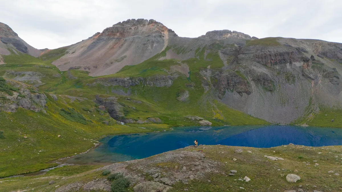 Turquoise alpine lake water fills the basin below steep gray cliffs and green tundra slopes beneath Mount Sneffels, with a few tiny hikers on the rocky foreground ridge. Soft morning light and a pale sky give the scene a calm, high-elevation feel.
