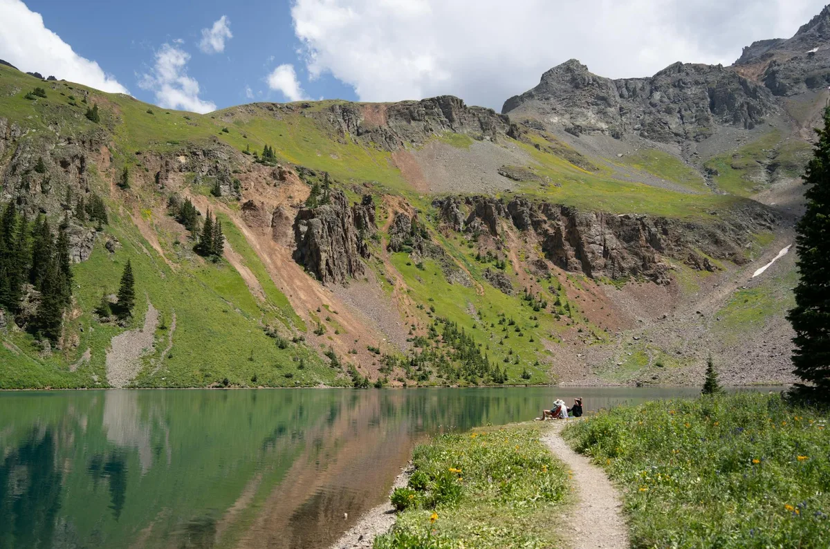 A turquoise alpine lake mirrors steep, rocky Mount Sneffels slopes under a bright morning sky, with a narrow trail and a few hikers in the wildflower-lined foreground. Green meadows and patches of summer snow add color against the gray cliffs and scree.